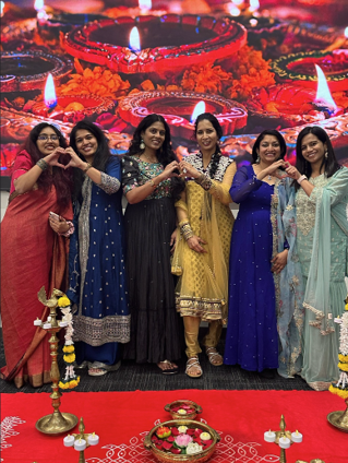 Group of women in colorful traditional attire posing in front of a Diwali-themed backdrop.