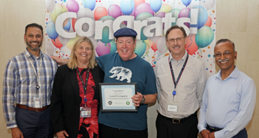 Five colleagues smiling, with one holding a certificate, standing in front of a colorful ‘Congrats’ balloon backdrop.
