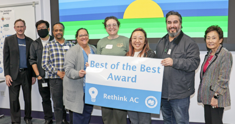 Eight people pose indoors holding a ‘Best of the Best Award – Rethink AC’ sign in front of a colorful presentation screen.