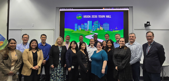 Group of colleagues posing in front of a ‘Vision 2036 Town Hall’ presentation screen.